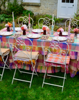 Outdoor table setting with a colorful plaid tablecloth and white chairs on a grassy area.