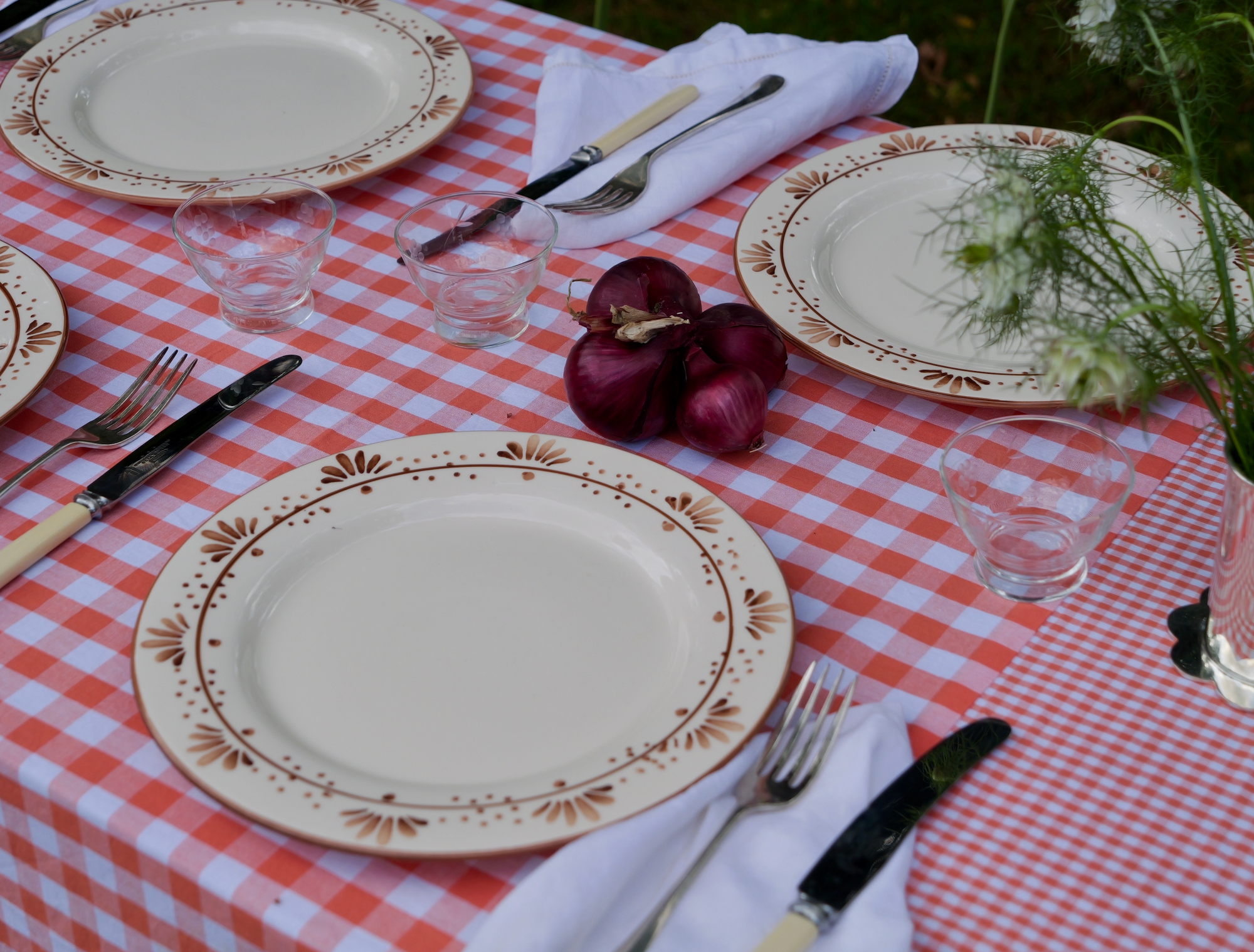 Outdoor dining table set with a hand-stitched orange patchwork gingham tablecloth, vintage floral china, white napkins, onions and red onions as decor, and a small vase of white wildflowers, surrounded by lush green garden foliage.