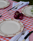 Outdoor dining table set with a hand-stitched orange patchwork gingham tablecloth, vintage floral china, white napkins, onions and red onions as decor, and a small vase of white wildflowers, surrounded by lush green garden foliage.