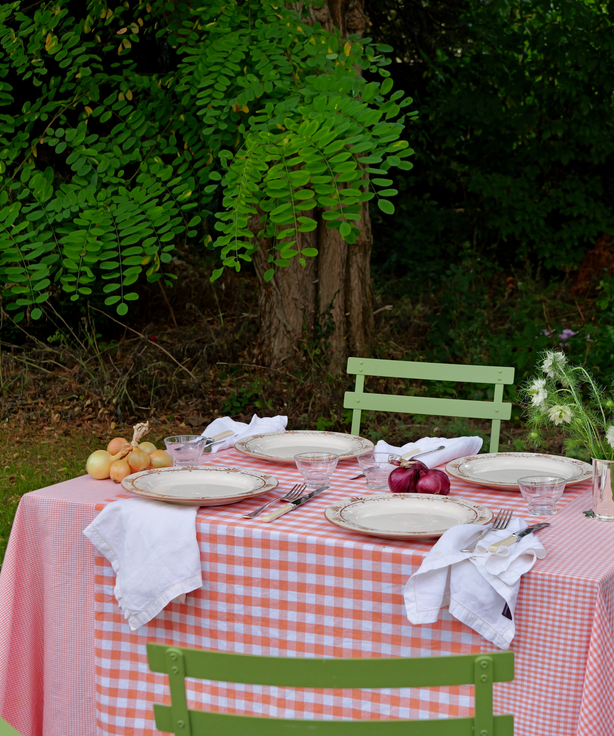 Outdoor dining table set with a hand-stitched orange patchwork gingham tablecloth, vintage floral china, white napkins, onions and red onions as decor, and a small vase of white wildflowers, surrounded by lush green garden foliage.