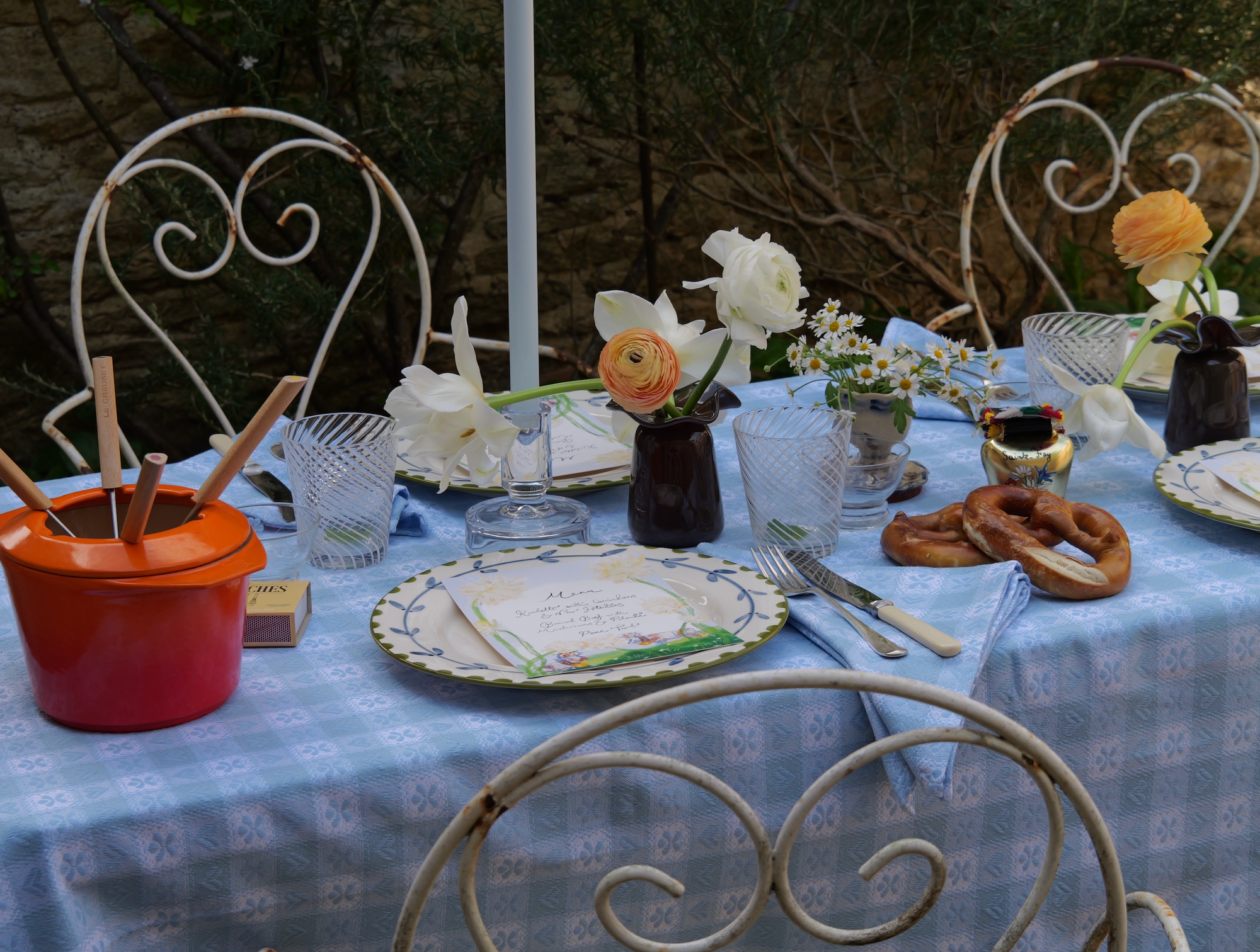 Rustic outdoor dining table with blue alpine tablecloth, white napkins, fresh flowers, and pretzels set against a stone wall.