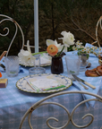 Rustic outdoor dining table with blue alpine tablecloth, white napkins, fresh flowers, and pretzels set against a stone wall.