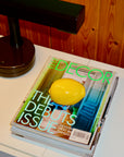 Decor magazine with a lemon on the cover on a table next to a lamp.