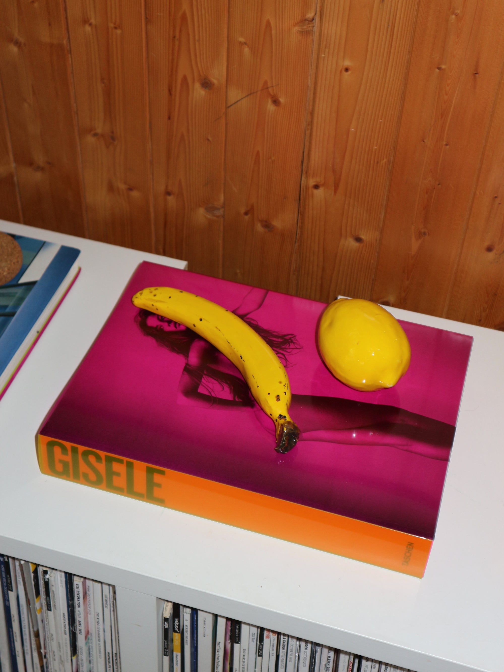 Book titled 'Gisele' with a banana and lemon on a pink surface against a wooden wall.