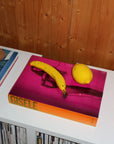 Book titled 'Gisele' with a banana and lemon on a pink surface against a wooden wall.